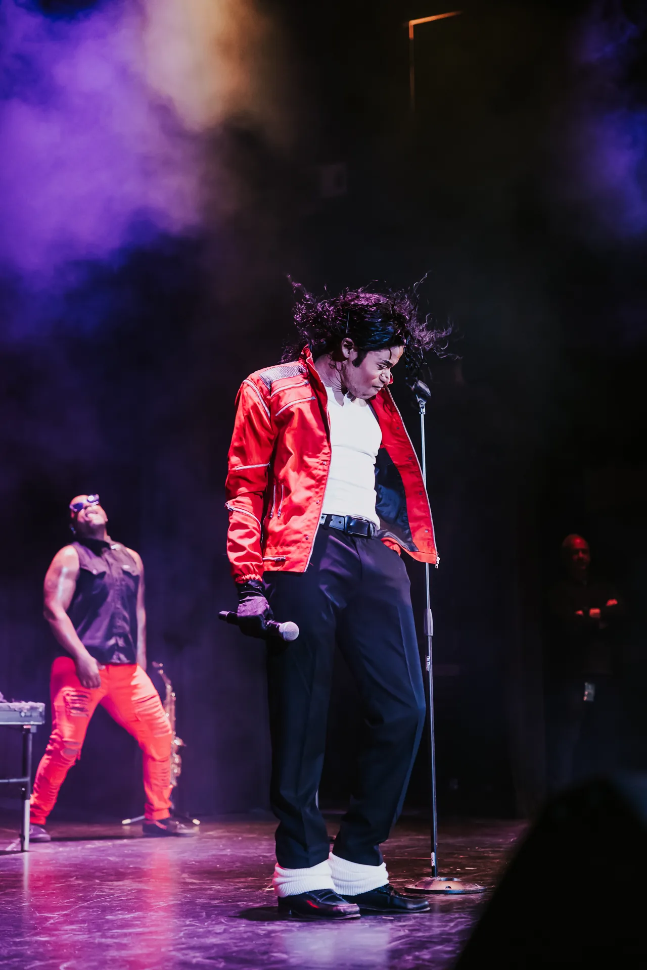Who’s Bad performer in a red jacket looks down beside a microphone stand as purple smoke fills the stage