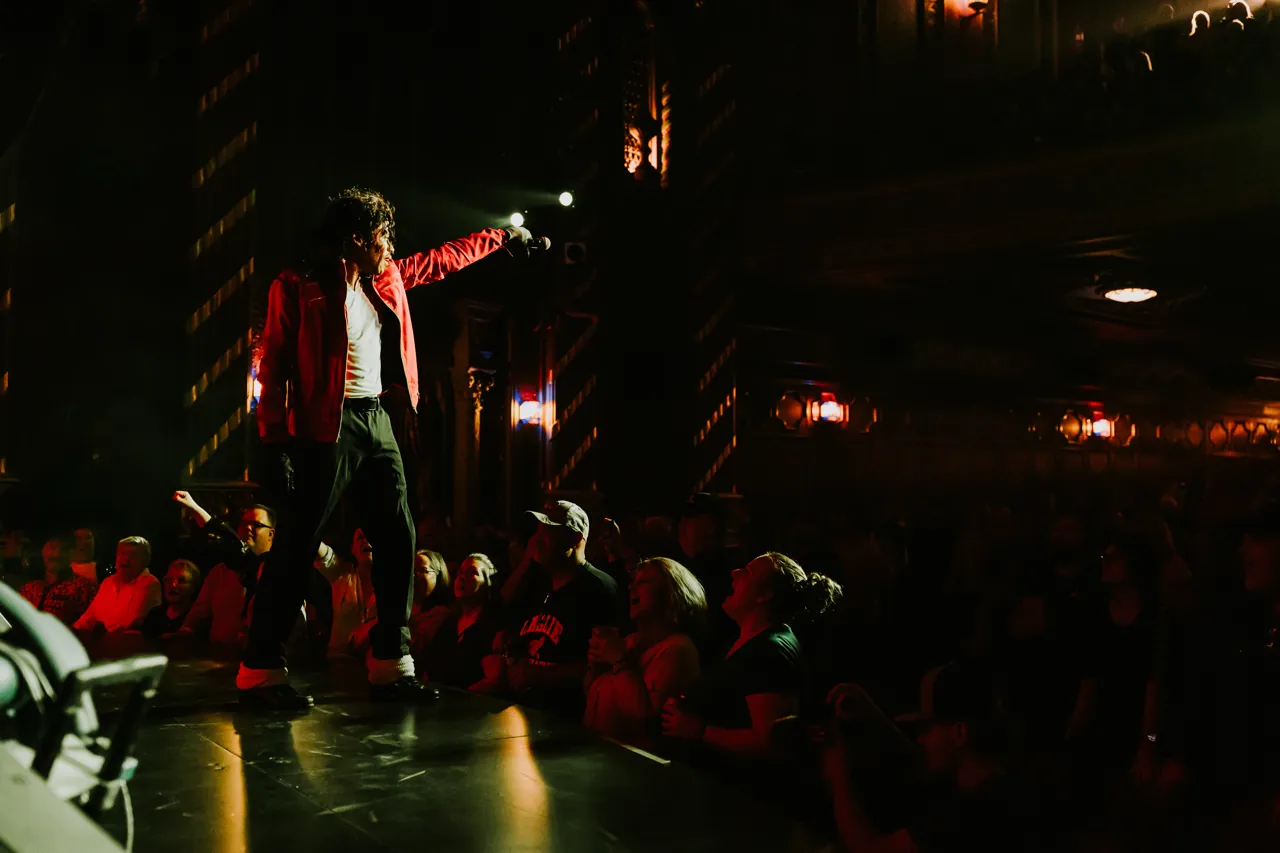 Who’s Bad performer in a red jacket points across a crowd of fans at the edge of the stage